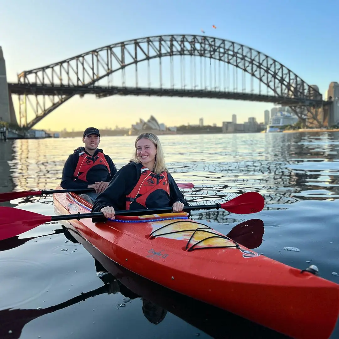 Girl and guy on a red canoe in Sydney Harbour with Harbour Bridge behind them on a sunny evening.