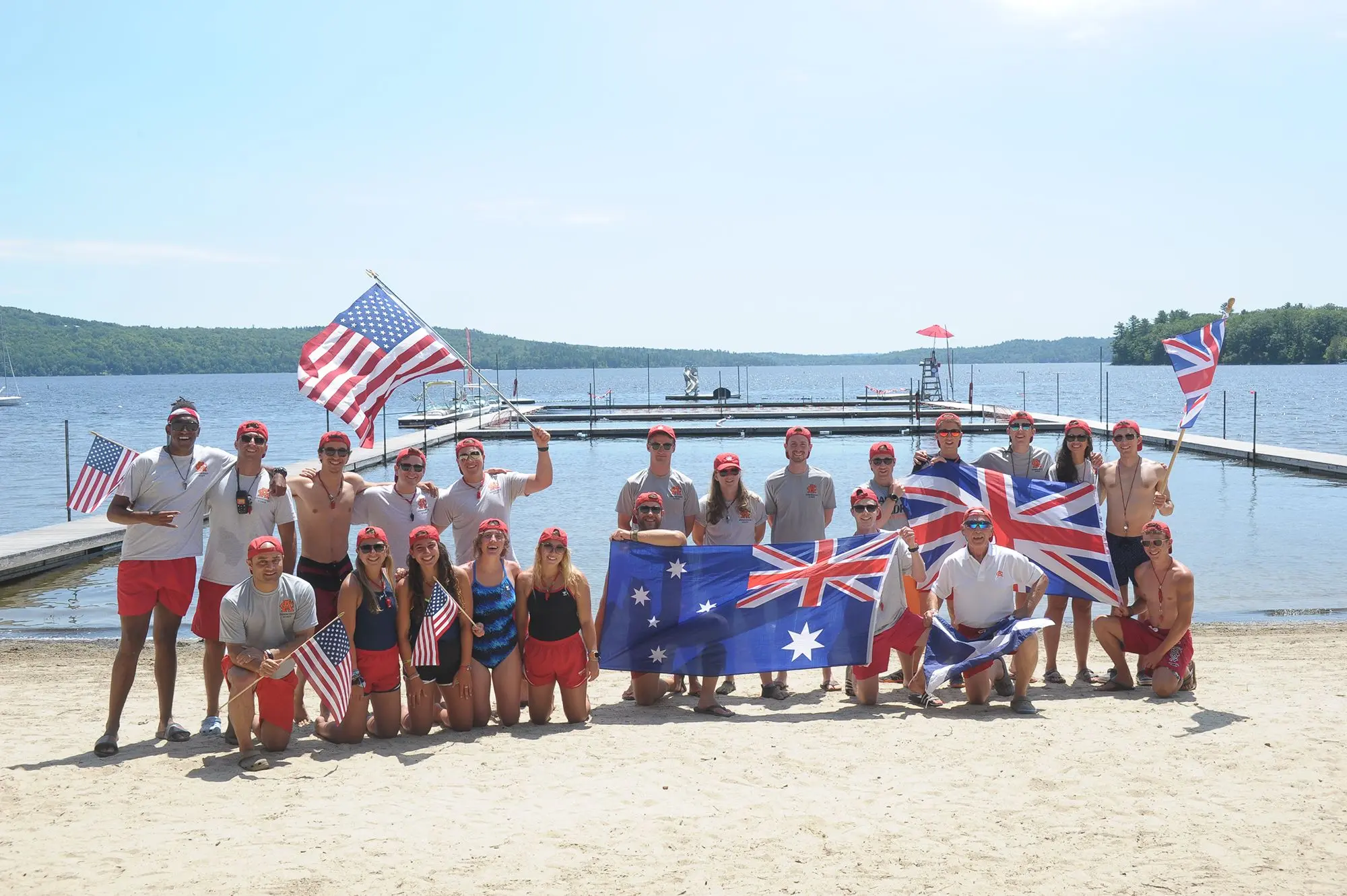 A group of campers stand in the sun next to a lake waving America, Australian and UK flags