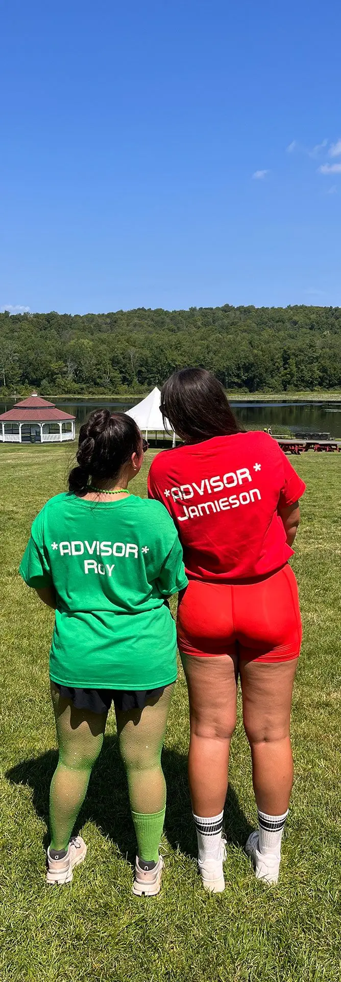 Two girls, one wearing red shorts and tshirt and one wearing green shorts and tshirt, facing away from camera on grass with lake and blue sky in the background