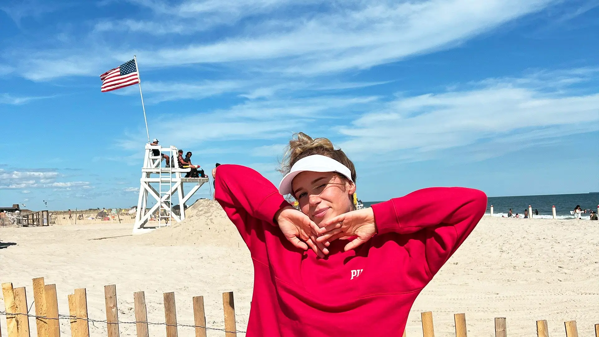 Blonde girl in red jumper on beach with American flag coming from lifeguard hut