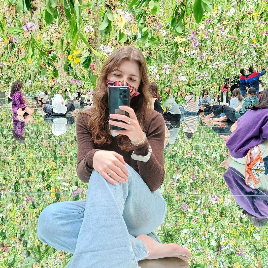 A young woman with brown hair and a facemask takes a selfie in a mirrored room full of flowers 