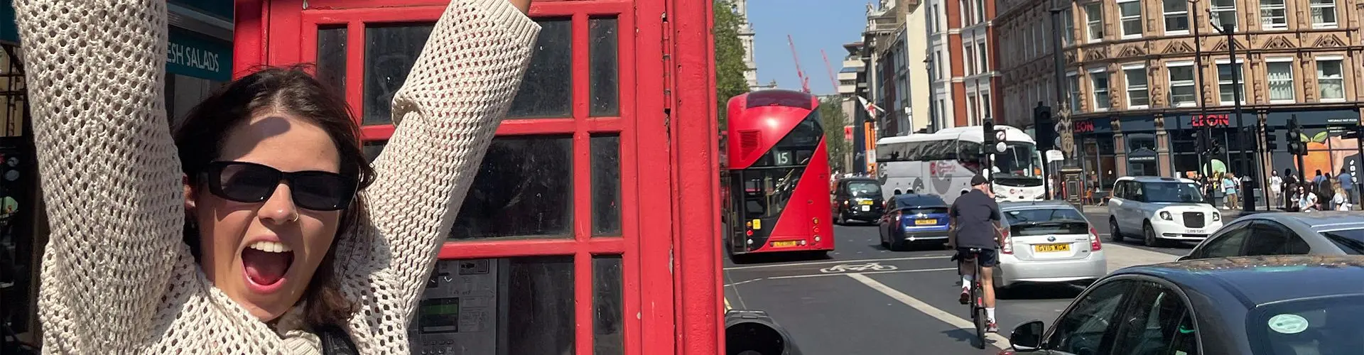 A young woman with sunglasses on and her hands in the air outside a phone box