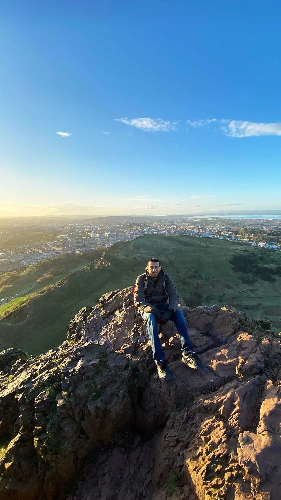 Guy sits on mountain beside Edinburgh on sunny morning.