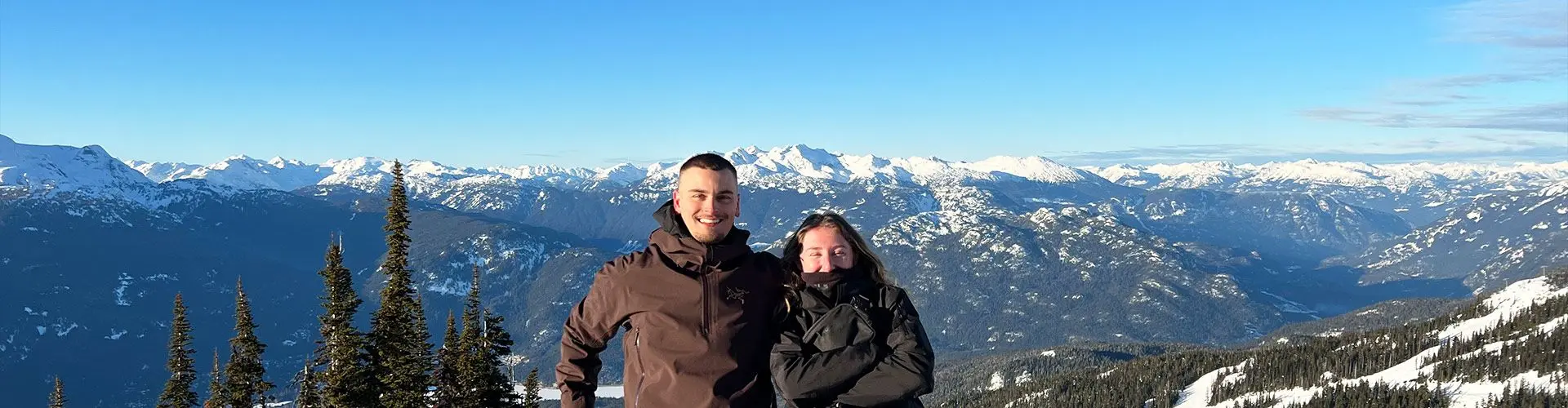 Guy and girl smile on snowy mountain with blue sky and mountains in background