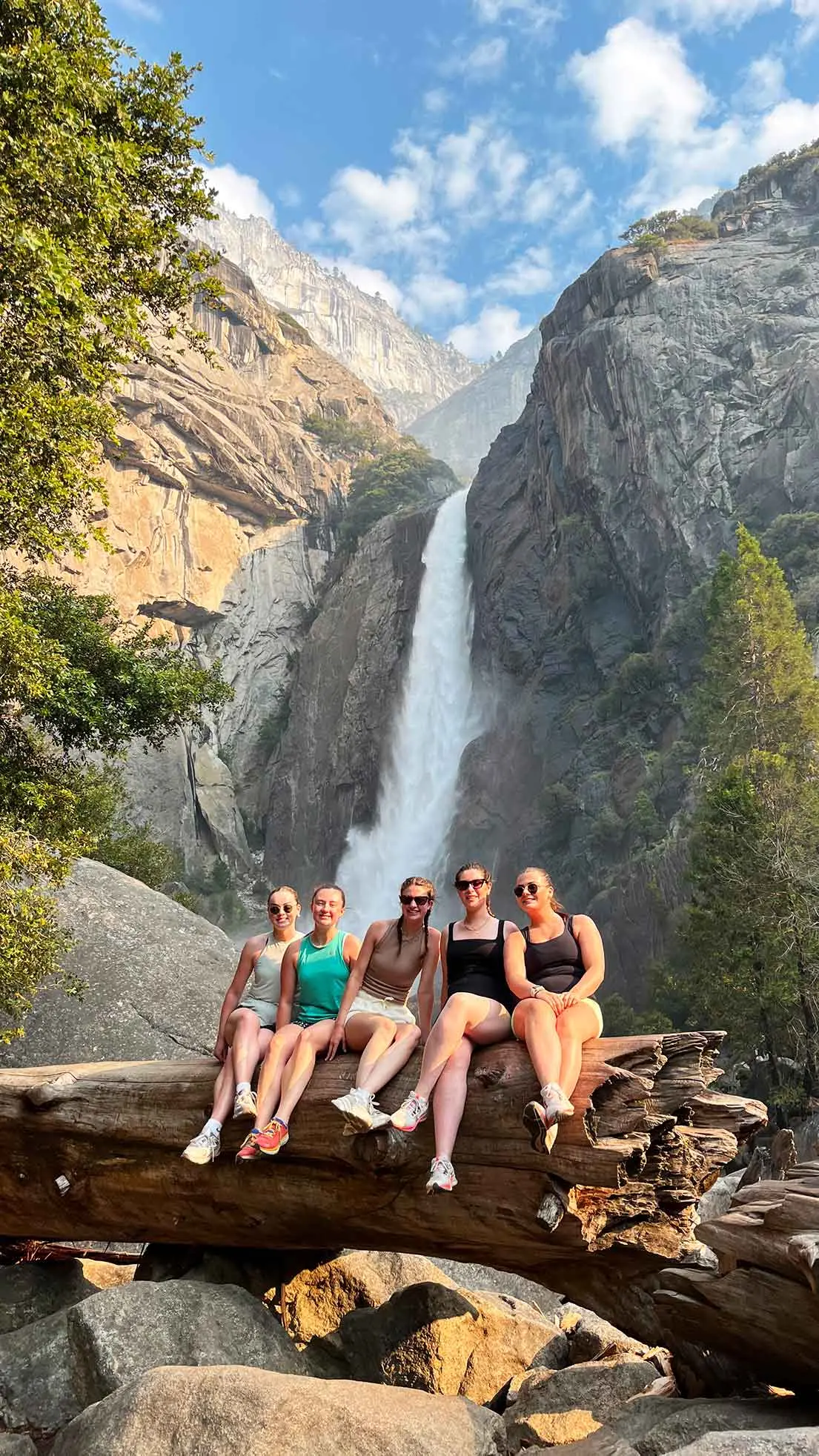 5 girls sit on fallen tree beneath waterfall.