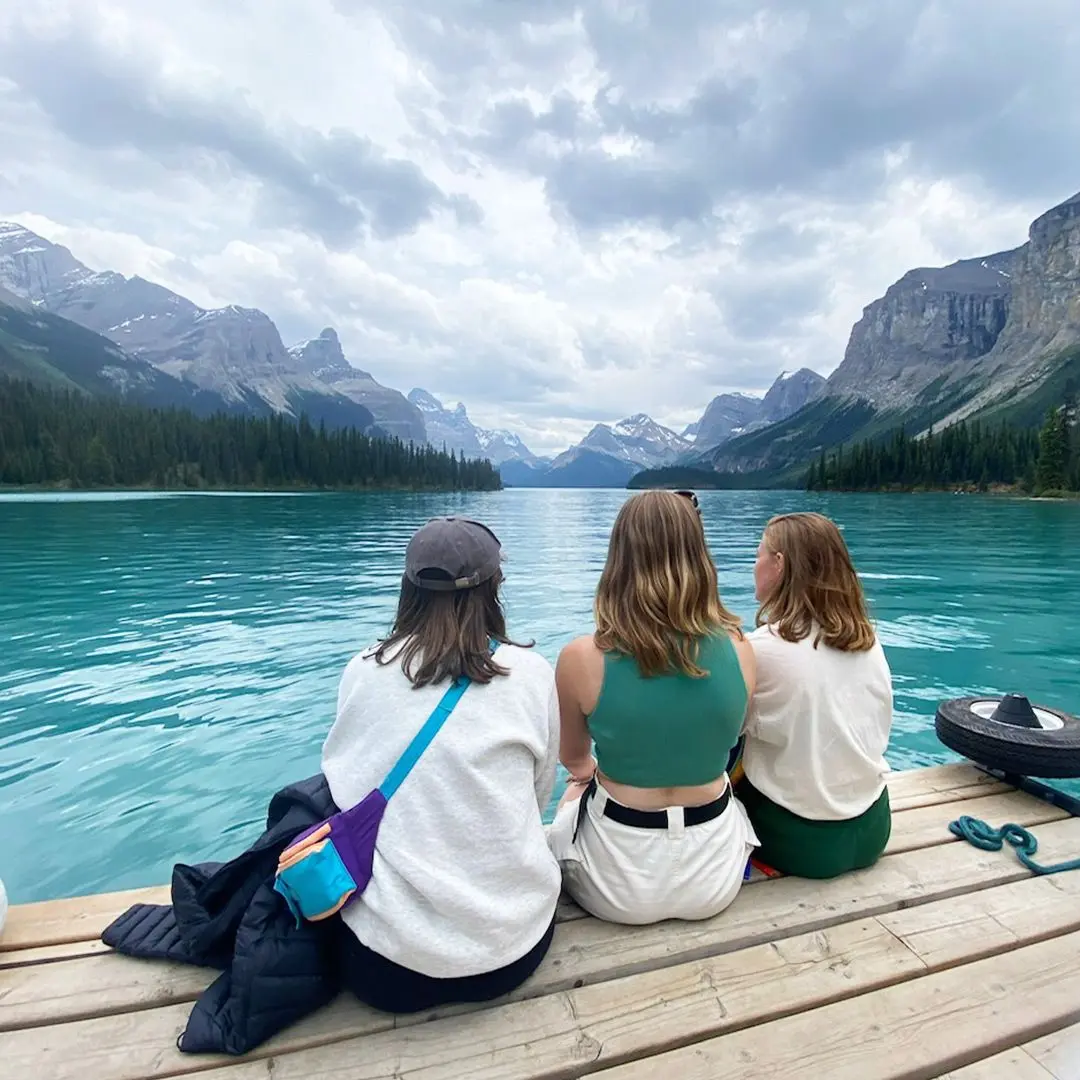 3 friends sit on edge of lake with mountains